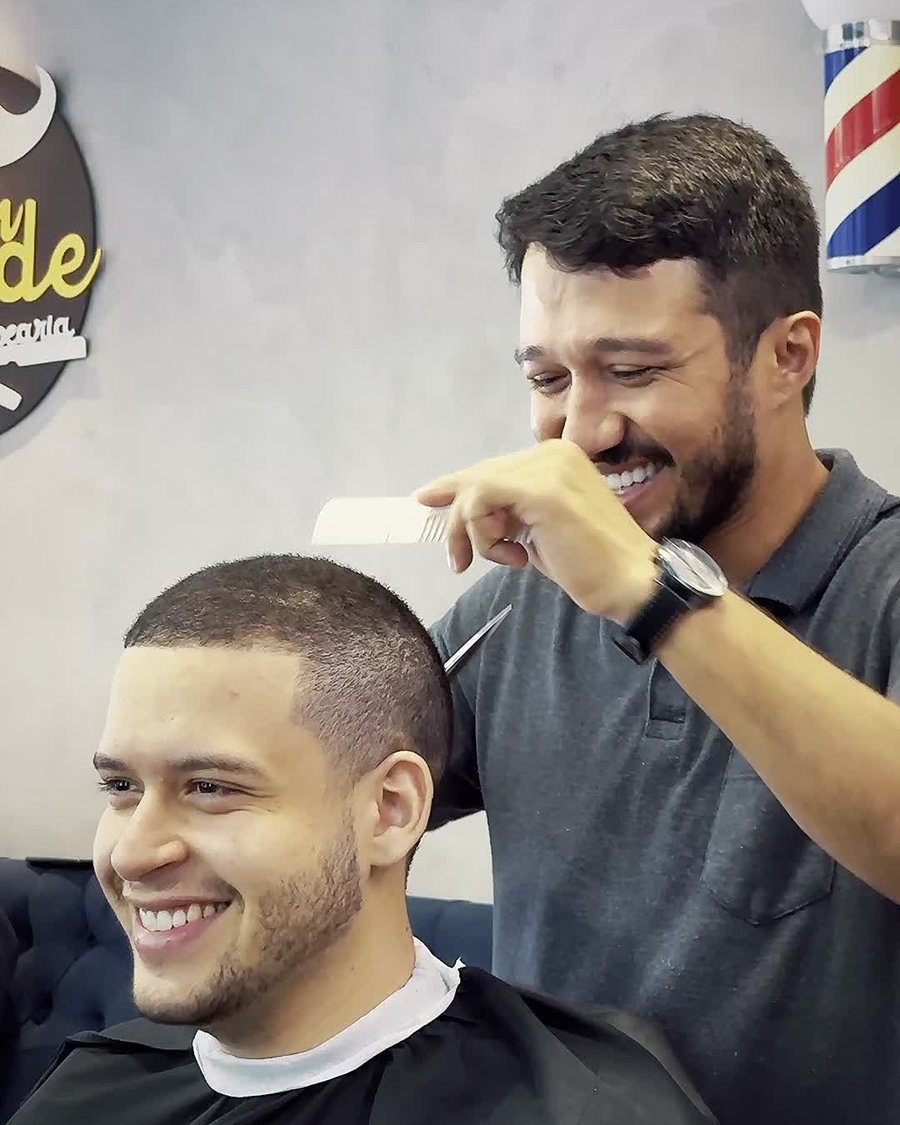 O barbeiro Manoel Gonçalves rindo com um cliente durante o corte, com o poste de barbeiro ao fundo